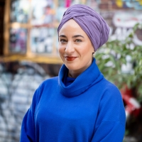Person mit Blauem Pullover und violetter Kopfbedeckung vor buntem Hintergrund